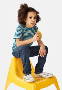 Barn med krøllet hår sidder på en gul plastikstol iført en blå og gråstribet skjorte, blå jeans og hvide sneakers, og holder en pæreformet drik.