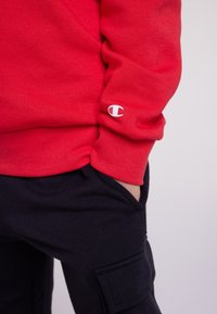 Persona che indossa una felpa rossa a maniche lunghe con il logo Champion sulla manica e pantaloni neri, con una mano nella tasca.