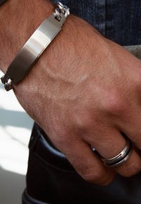 Zilveren armband met een platte metalen oppervlakte en een gevlochten ontwerp, gedragen aan een hand met een zilveren ring om een andere vinger. Gestructureerde huid zichtbaar.