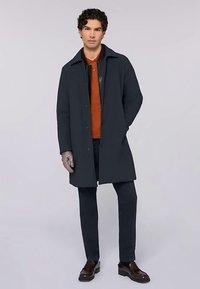 Ciemnoniebieski płaszcz z wyłogiem z wcięciem, noszony na pomarańczowej koszuli. Model ma na sobie szare rękawiczki i ciemne spodnie oraz brązowe buty.