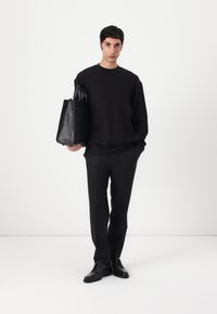 Sweatshirt noir à coupe décontractée, associé à un pantalon noir slim et des chaussures noires brillantes. Un sac fourre-tout en cuir noir est tenu sur le côté.