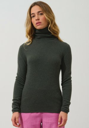 Pull à col roulé vert foncé en matériau doux, au design ajusté avec des manches longues, associé à un pantalon rose. Fini simple et texturé.