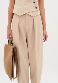 Top beige senza maniche con bottoni neri, abbinato a pantaloni beige a vita alta e una borsa a tracolla in pelle color tan, caratterizzati da texture lisce.