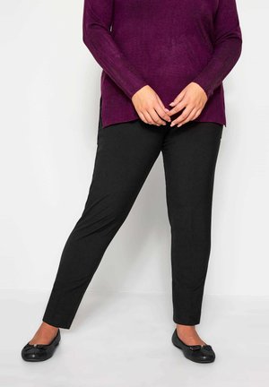 Personne portant un pantalon noir, des ballerines noires et un pull violet à manches longues, debout les mains jointes devant.
