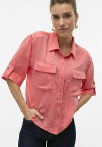 Camicia a manica corta rosa corallo con bottoni e due taschini sul petto. Realizzata in tessuto leggero, presenta una vestibilità comoda e maniche arrotolate.