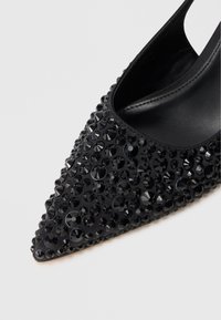 Pantof cu vârful ascuțit, negru, decorat cu strasuri negre dense pe partea superioară, având un lateral tăiat jos și o căptușeală interioară netedă.
