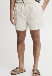 Pantalones cortos de algodón beige con cintura elástica y cordón. Presentan bolsillos laterales y un ajuste relajado, combinados con sandalias marrones.