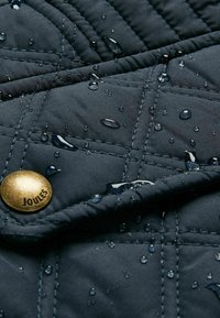 Quiltelt sötétkék szövet vízcseppekkel, egy "JOULES" felirattal gravírozott arany gombbal, kiemelve a nedvességállóságot.