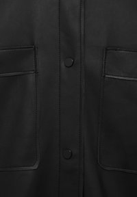 Gros plan d’un vêtement en cuir noir avec deux poches rectangulaires à l’avant et deux boutons-pression noirs sur la couture centrale avant.