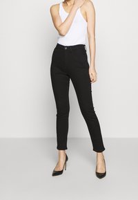 Mujer vestida con pantalones ajustados negros, top sin mangas blanco y zapatos de tacón alto negros, de pie con una mano cerca de su hombro.