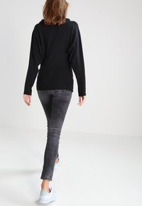 Maglione nero oversize con spalle scese, abbinato a jeans grigio chiaro. La calzatura include sneakers bianche. Sfondo minimalista.