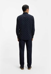 Camicia blu navy con piccolo motivo triangolare bianco, maniche lunghe e colletto button-down, abbinata a pantaloni di denim scuro e scarpe marroni.