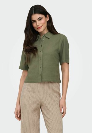 Femme aux longs cheveux noirs portant une chemise kaki à manches courtes boutonnée et un pantalon beige texturé, debout devant un fond clair.