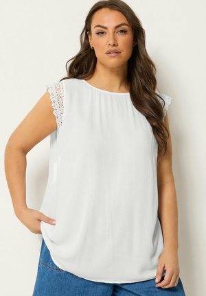 Kvinde med langt brunt hår iført en hvid ærmeløs bluse med blondedetaljer og blå jeans, stående med den ene hånd i lommen.