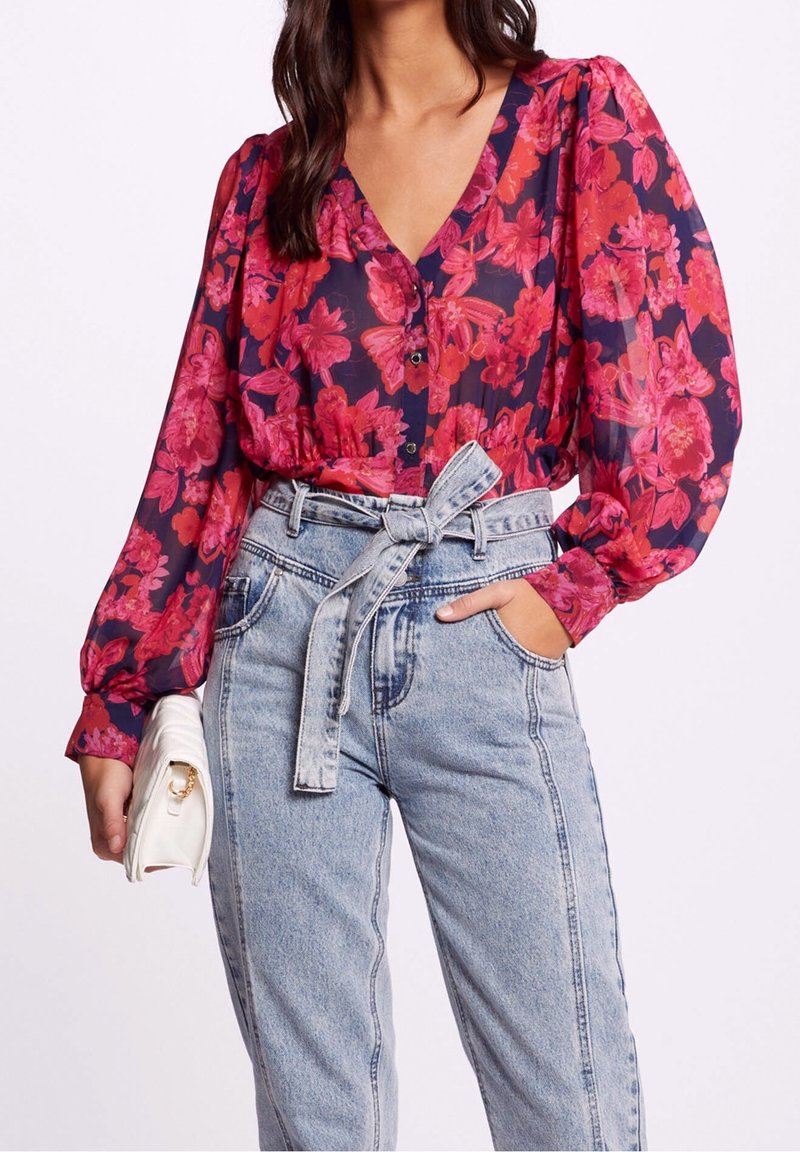 Frau trägt eine langärmlige rot-rosa gefärbte Bluse mit Blumenmuster, die in hellblaue hochgerückte Jeans mit gebundenem Gürtel gesteckt ist, während sie eine weiße Clutch hält.