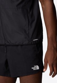 Black windbreaker with a smooth texture, featuring a zip closure and the "WINDWALL" label. Paired with black, textured shorts and logo detail.