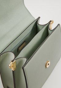 Zelená kožená kabelka s hladkým povrchem, zdobená zlatými kovovými prvky a dvěma přihrádkami. Uzávěr obsahuje kulatý kovový tlačítko.