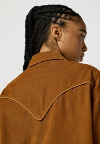 Chemise en velours côtelé de couleur marron riche avec un col et un détail de pli incurvé. Équipée de rayures verticales et d'une texture douce.