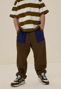 Gestreept terrycloth kortlossen shirt in olijfgroen en wit, gedragen met bruine loszittende broek met blauwe zijzakken. Gemaakt met een paar meerkleurige sneakers.