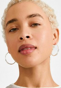 Lähivõte inimesest, kellel on lokkis blondid juuksed ja ta kannab hõbedaseid rõngakujulisi kõrvarõngaid. Iseloomustavad omadused on sile nahk, väljendunud huuled ja loomulik meik.