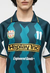 Νέος φοράει ποδοσφαιρική φανέλα με πράσινες και τυρκουάζ ρίγες, με το λογότυπο Replay Inc, τον αριθμό 11 και το κείμενο "Engineered Goods" στο μπροστινό μέρος.