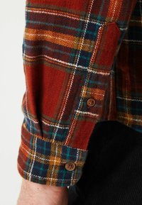 Chemise en flanelle à carreaux rouges avec des accents bleu marine, vert et marron, dotée d'un poignet à bouton et d'un tissu texturé. Comprend des détails de couture blanche.