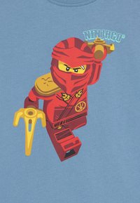 Sarkana ninjas grafika gaišā zilos t-kreklā, ar zelta ieročiem un melniem akcentiem, un tekstu "Ninjago" augšpusē.