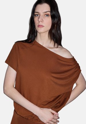 Junge Frau mit langen dunklen Haaren trägt ein braunes schulterfreies Oberteil und posiert mit einer Hand auf der Hüfte vor einem schlichten Hintergrund.