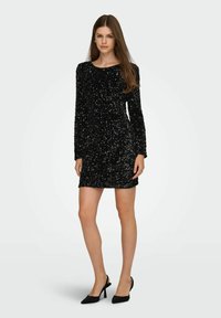 Vestido negro de lentejuelas con mangas largas y escote redondo; silueta ajustada, largo hasta la rodilla, combinado con zapatos de tacón negro.
