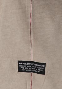 Tela beige con costuras rojas prominentes a lo largo de una costura. Presenta una etiqueta negra con texto blanco, detallando la marca y el eslogan.