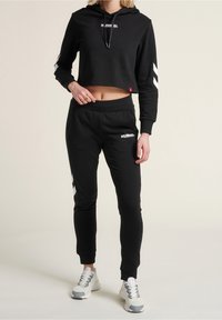 Felpa nera corta con logo "HUMMEL", abbinata a pantaloni della tuta neri con strisce laterali e logo. Cotone testurizzato, vestibilità rilassata.