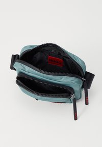 Geantă crossbody din țesătură teal, cu două compartimente cu fermoar, având interior negru, etichetă roșie a mărcii și accesorii contrastante de culoare neagră.