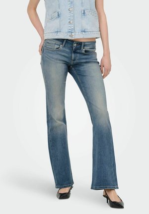 Kobieta ubrana w wyblakłe niebieskie dżinsy typu bootcut, jasnoniebieską bezrękawnikową kamizelkę dżinsową oraz czarne, spiczaste szpilki z paskami na kostce.