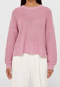 Femme portant un pull en maille rose ample à manches longues et un pantalon blanc taille haute à jambes larges.