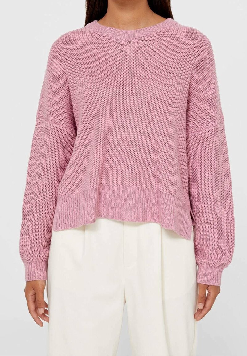 Femme portant un pull en maille rose ample à manches longues et un pantalon blanc taille haute à jambes larges.
