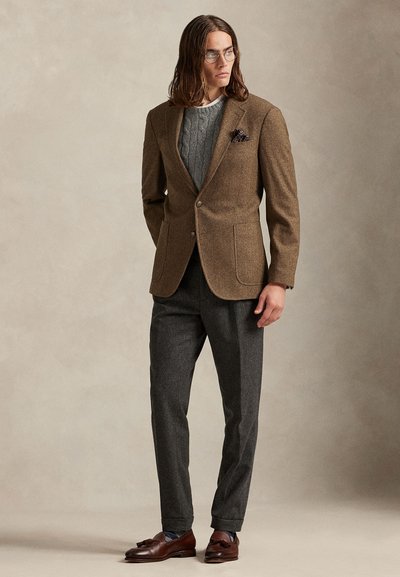 Άνδρας με μαλλιά μέχρι τους ώμους που φοράει γυαλιά, καφέ σακάκι, γκρι πουλόβερ με κοτσίδες, γκρι παντελόνι και καφέ μοκασίνια με φούντες, στέκεται μπροστά σε μπεζ τοίχο.