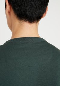 Ciemnozielona bawełniana koszulka z okrągłym dekoltem ma gładkie, teksturowane wykończenie z subtelnymi szwami w okolicy dekoltu i ramion.