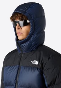 Puffjacka med marinblå huva, svart kropp, kviltad yta och dragkedja fram. Har "The North Face"-logotypen på bröstet.