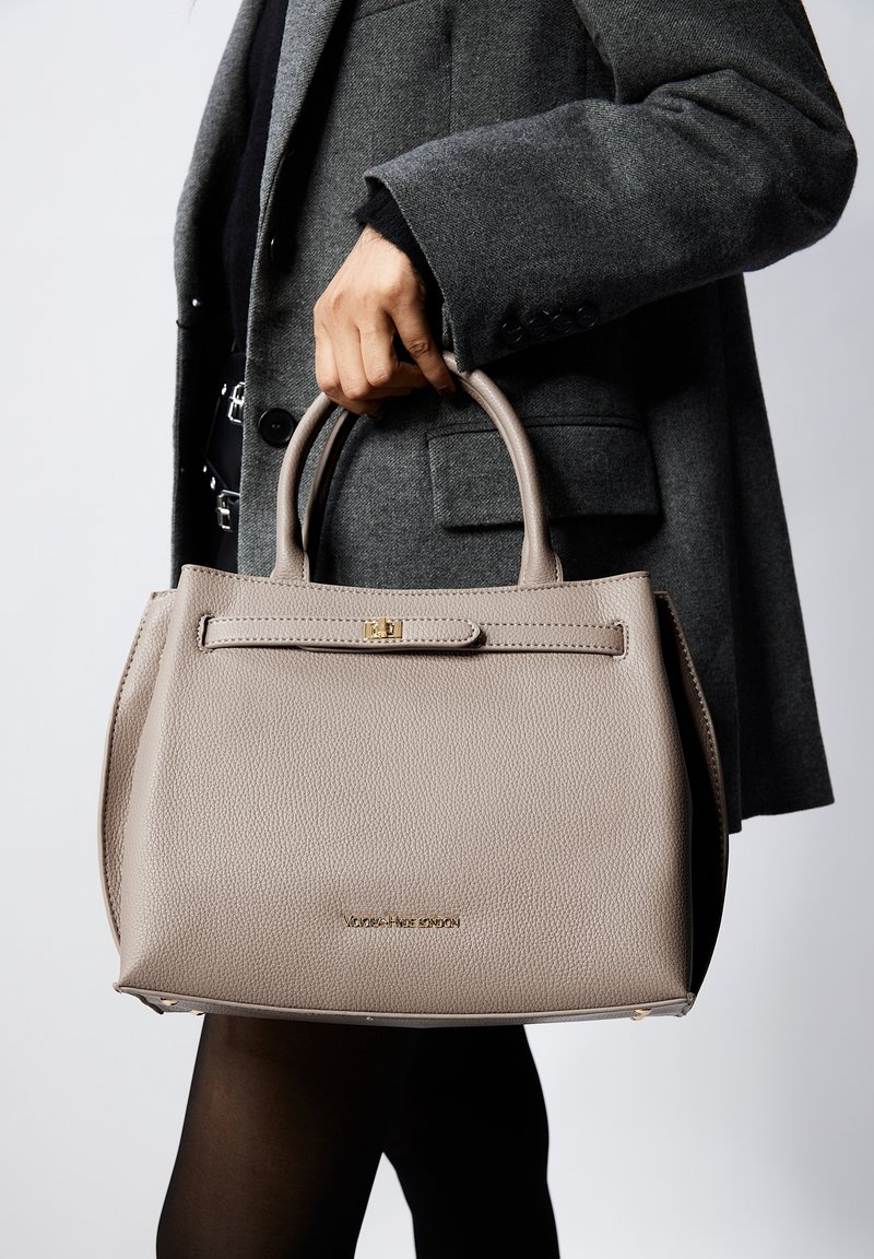 Sac à main en cuir beige avec un design structuré, des poignées supérieures et un fermoir doré. Présente un matériau texturé et un logo de marque discret en bas.