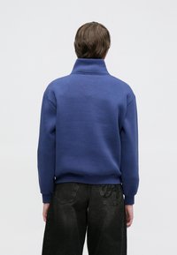 Tumši zils pulovers ar augstu apkakli, ribots aproces un apakšmala. Tam ir gluda tekstūra un brīvs piegriezums.