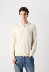 Cream long-sleeve polo shirt with a zip collar and black "BOSS" logo. Features ribbed cuffs and a relaxed fit. Worn with blue jeans.