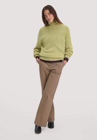 Hellgrüner, gestrickter Rollkragenpullover mit gerippten Bündchen, kombiniert mit braunen, weit geschnittenen Hosen und schwarzen Stiefeletten. Einfarbiger Hintergrund.