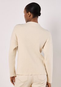 Beige gerippter Pullover mit hohem Kragen, langen Ärmeln und einer lockeren Passform, der dezente Seitendetails für Textur und Flexibilität aufweist.