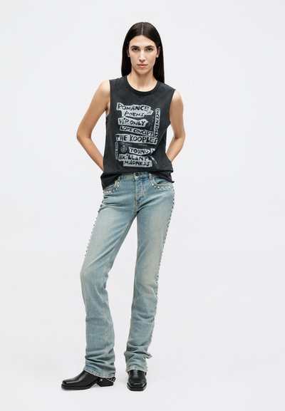 Woman stands with hands behind back, wearing sleeveless black graphic top and light blue jeans with studded side details and black boots.