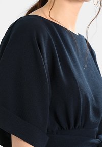Granatowa, strukturalna bluzka z okrągłym dekoltem i szerokimi krótkimi rękawami, z marszczeniami w talii. Miękka w dotyku tkanina.