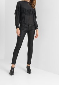 Top negro de manga larga con acentos de volantes, combinado con jeans ajustados negros y botines de tacón negro, sobre un fondo neutro.