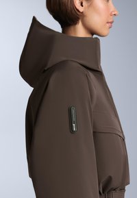 Hnedá nepremokavá bunda s vysokým golierom, hladkou textúrou a štítkom RECCO na ľavom rukáve. Detaily švov zdôrazňujú dizajn.