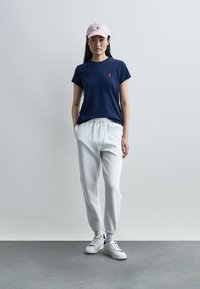 Νεαρή γυναίκα που στέκεται, φορώντας μπλε σκούρο μπλουζάκι, λευκά φούτερ παντελόνια, λευκά αθλητικά παπούτσια και ροζ ανοιχτό baseball καπέλο, σε απλό φόντο.