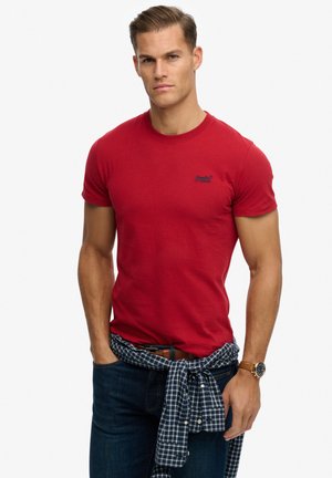 Mand iført en tætsiddende rød t-shirt, mørke jeans, en ternet skjorte bundet om livet og et armbåndsur, stående med hænderne i lommerne.