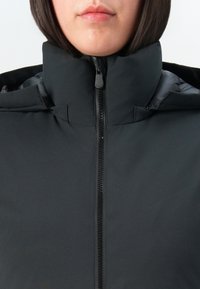 Femme portant une veste matelassée noire avec un col montant fermé par une fermeture éclair, visible uniquement de la bouche jusqu'à la poitrine supérieure, sur un fond uni.